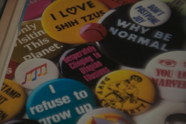 Blurry photo of a pinback button that says 'Desperately Clinging to Utopian Illusions' as well as several more buttons.